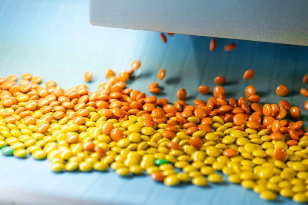 W fabryce w Janaszówku zaawansowana technologia wspiera, a nie zastępuje ludzi Kolorowe tabletki w odcieniach żółtego i pomarańczowego przesypujące się na taśmociąg na linii produkcyjnej w fabryce farmaceutycznej.