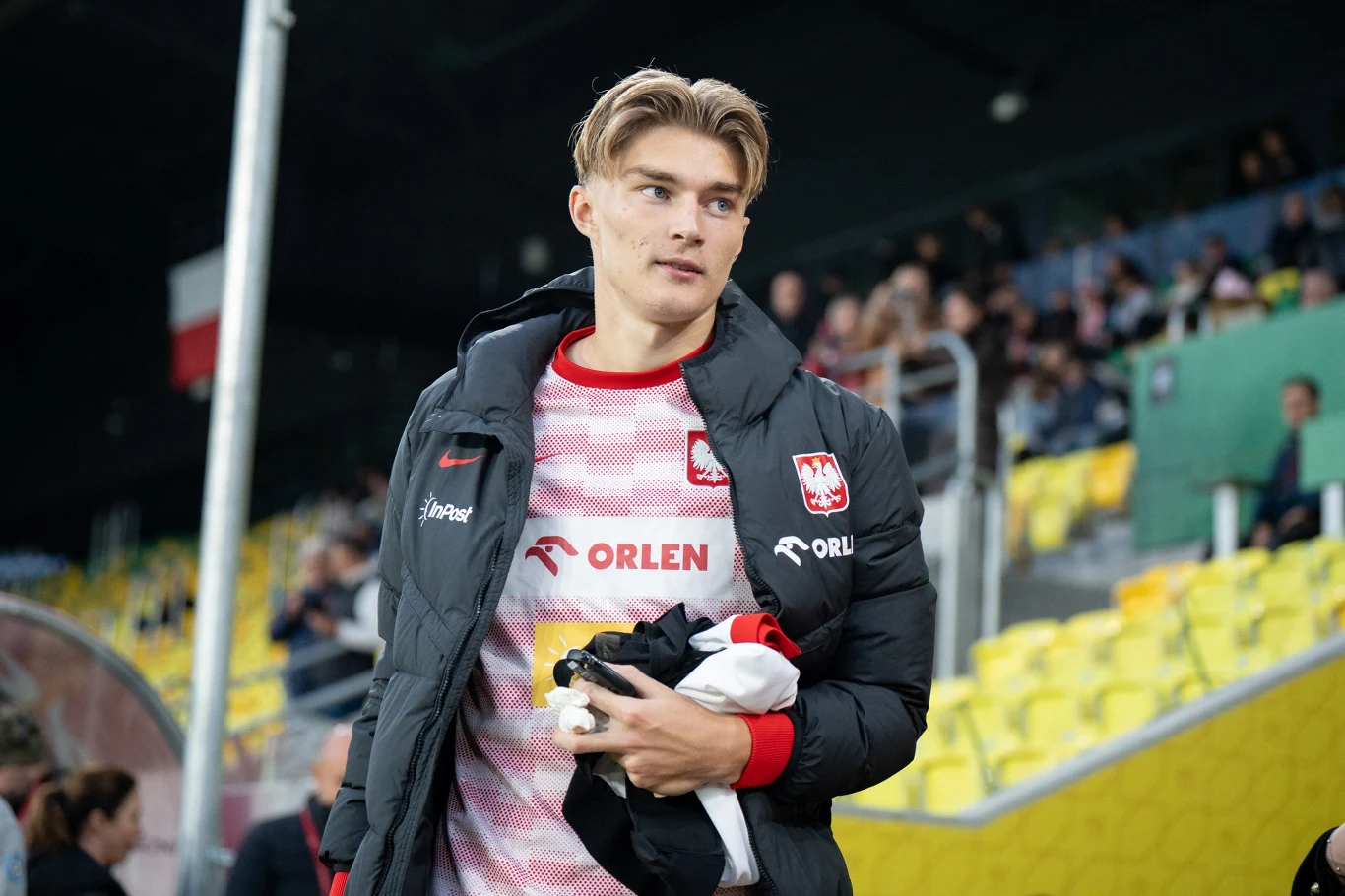 Młody mężczyzna w sportowej kurtce i koszulce z emblematem reprezentacji Polski stoi na stadionie piłkarskim, trzymając w rękach ubrania. W tle widoczne żółte siedzenia oraz grupa kibiców.