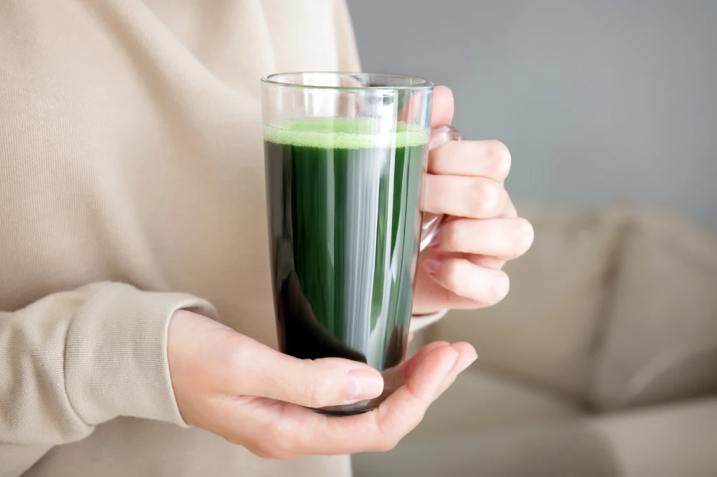 Jedna szklanka dziennie zamiast kawy. Daje energię i oczyszcza organizm Szklanka z zielonym napojem trzymana w dwóch dłoniach przez osobę ubraną w beżowy sweter, tło rozmyte w neutralnych odcieniach.
