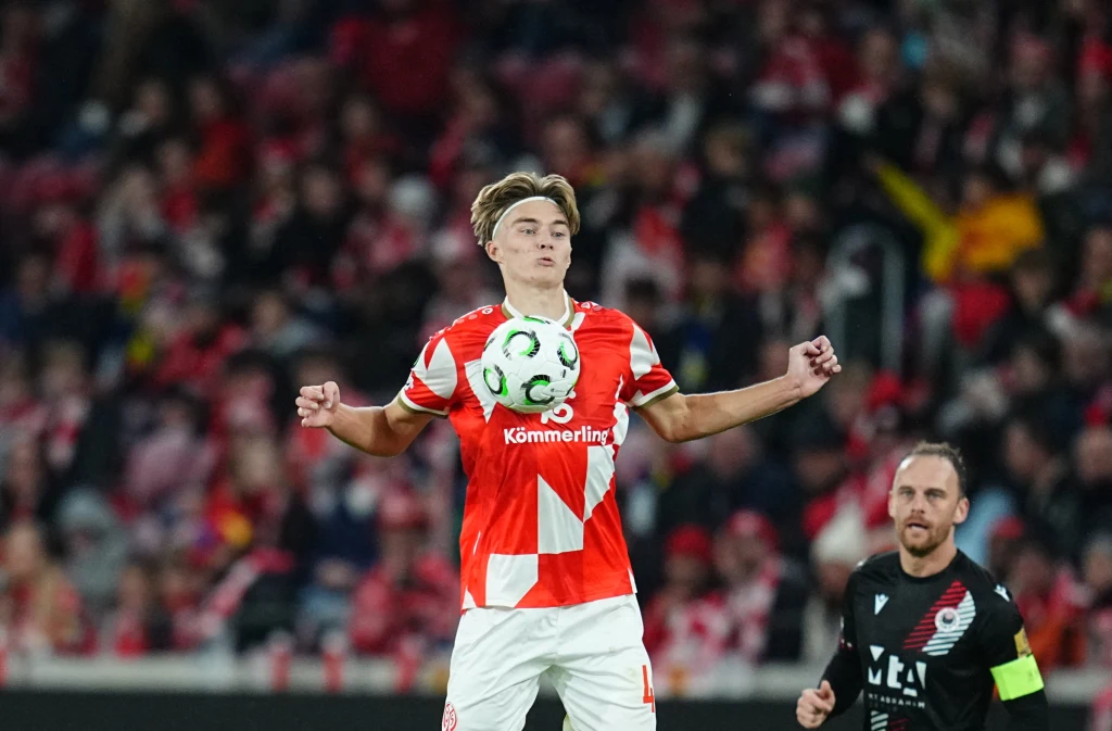 Kacper Potulski Zawodnik w czerwono-białej koszulce kontroluje piłkę klatką piersiową podczas meczu piłkarskiego, w tle widoczna grupa kibiców na trybunach oraz inny zawodnik z opaską kapitana.