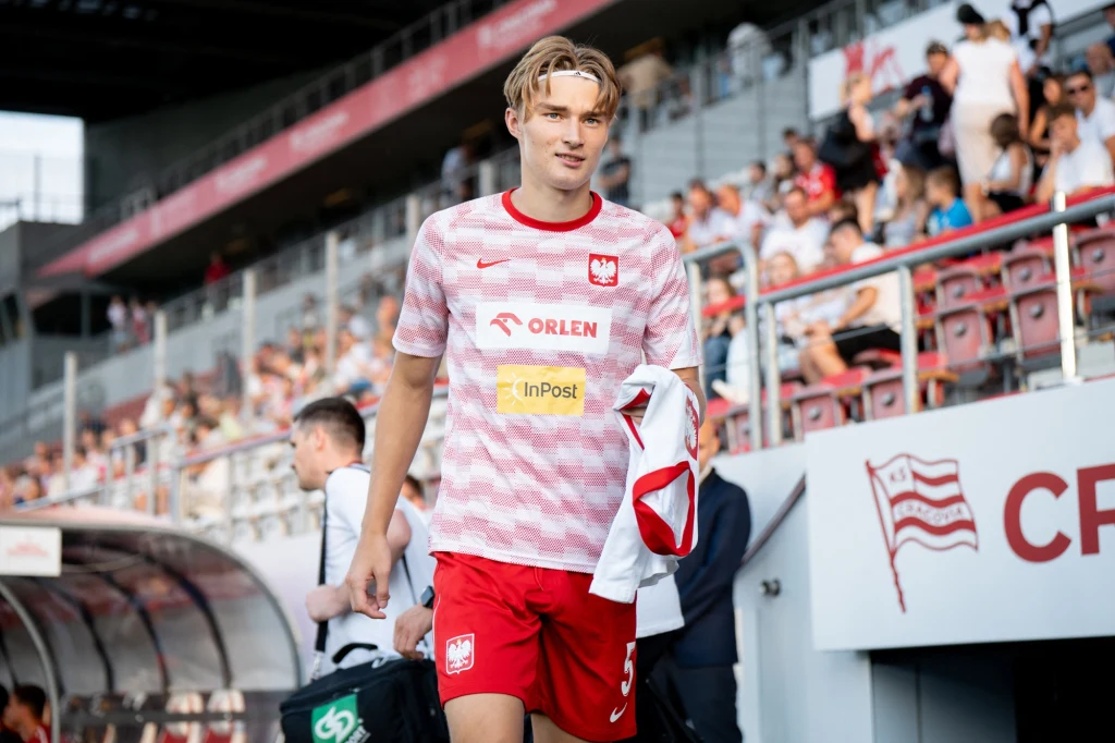 Kacper Potulski Młody piłkarz w stroju reprezentacji Polski opuszcza boisko, w tle trybuny z kibicami oraz elementy stadionu, widoczne logo sponsorów na koszulce zawodnika.