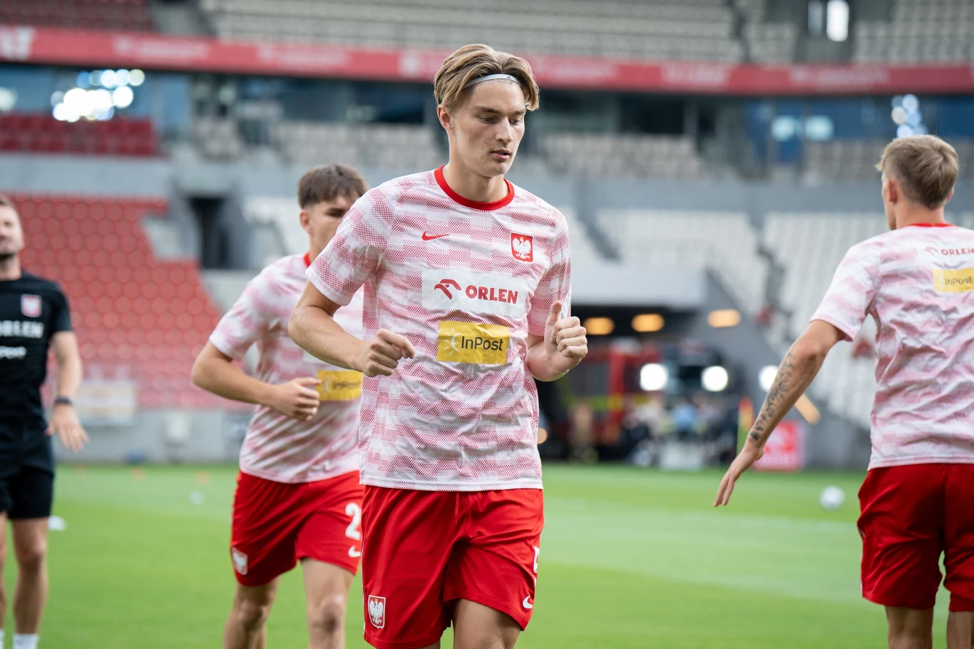 Kacper Potulski Piłkarze w biało-czerwonych strojach treningowych z logo Orlen i InPost biegną na stadionie, na pierwszym planie zawodnik z dłuższymi blond włosami, wokół nich rozciąga się trybuna stadionowa z pustymi miejscami.