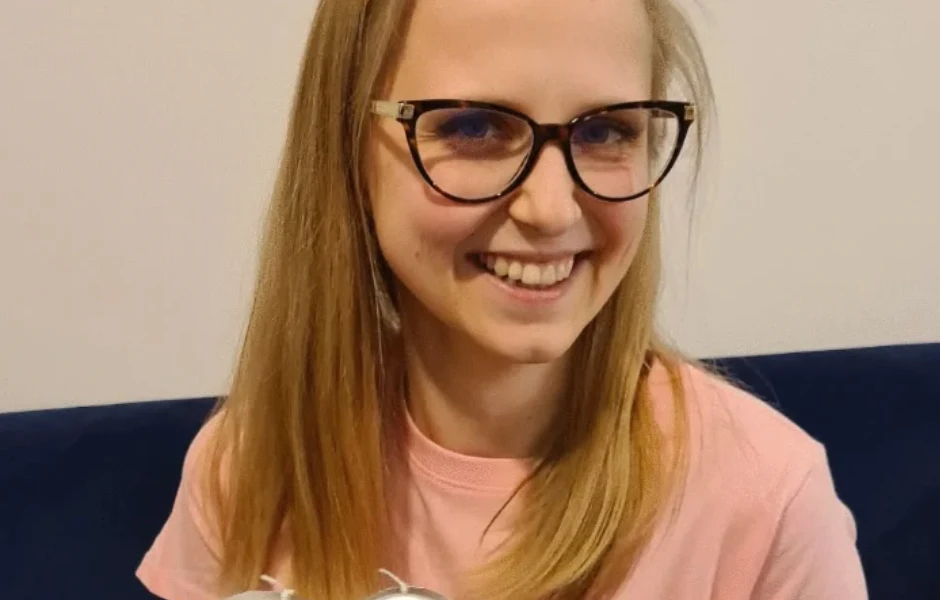 "Ślub od pierwszego wejrzenia": Agnieszka Miezianko spełniła marzenia Młoda kobieta w okularach z długimi blond włosami, uśmiechnięta, ubrana w różową koszulkę, siedzi na tle niebieskiej kanapy.