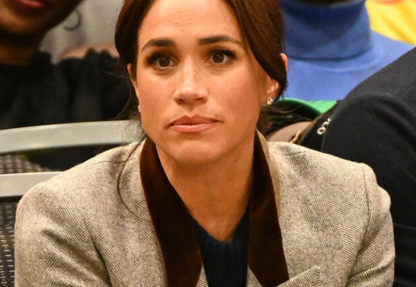 Meghan Markle Kobieta o ciemnych włosach spiętych do tyłu w eleganckiej, jasnej marynarce z ciemnobrązowym kołnierzem siedzi wśród innych osób, patrząc poważnie przed siebie.