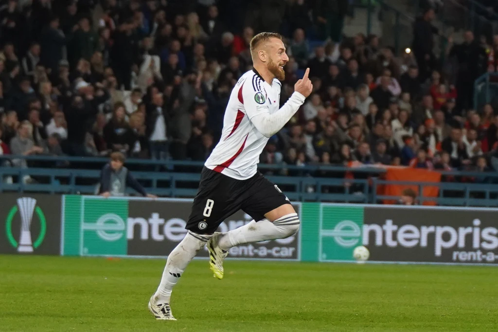 Rafał Augustyniak Piłkarz w białym stroju z numerem 8 biegnie po boisku, unosząc zaciśniętą pięść i podnosząc jeden palec do góry, w tle trybuny wypełnione kibicami.