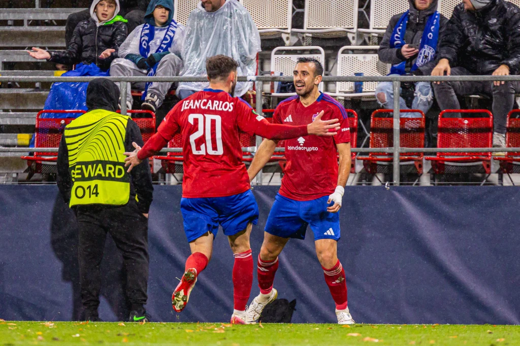 Zawodnicy Rakowa Częstochowa Dwóch piłkarzy w czerwonych koszulkach cieszy się po zdobyciu bramki na stadionie, w tle kibice i steward w żółtej kamizelce, widoczne mokre siedziska i kurtki sugerujące deszczową pogodę.