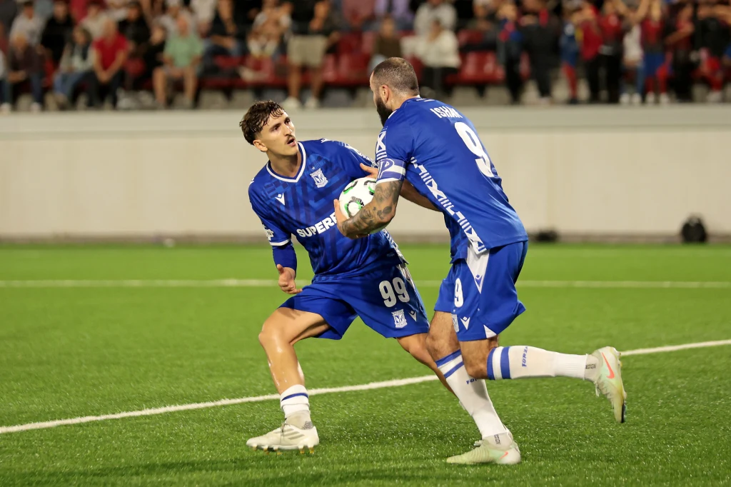 Lech Poznań zanotował upokarzającą porażkę z Lincoln Red Imps Dwóch piłkarzy w niebieskich strojach na boisku świętuje zdobycie gola, jeden z nich dynamicznie krzyczy z uniesioną pięścią, a drugi trzyma piłkę, wokół widownia na trybunach.