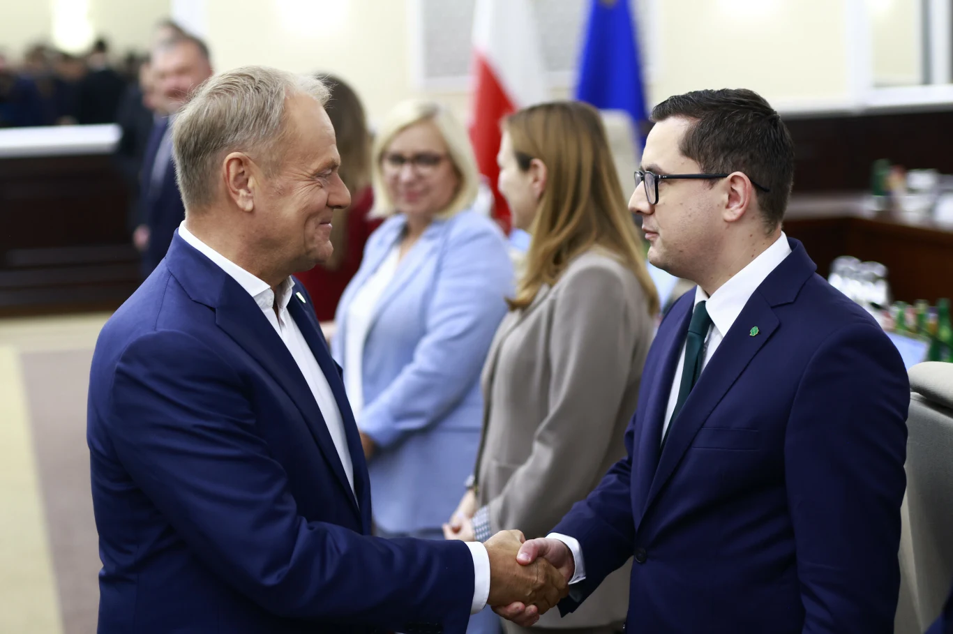 Dwóch mężczyzn w garniturach wymienia uścisk dłoni w oficjalnym wnętrzu, w tle trzy osoby w formalnych strojach oraz widoczne flagi Polski i Unii Europejskiej.