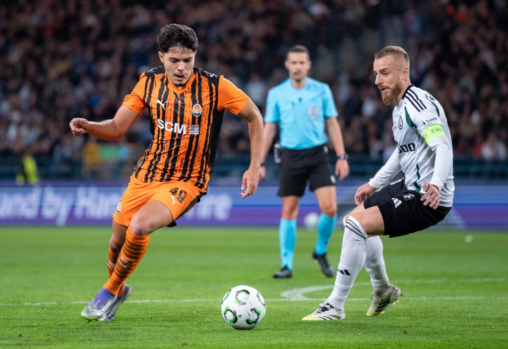 Rafał Augustyniak w meczu z Szachtarem Donieck Dwóch piłkarzy rywalizujących o piłkę podczas meczu piłkarskiego na stadionie, w tle sędzia oraz widownia. Jeden z zawodników nosi strój w pomarańczowo-czarnych pasach, drugi biały strój z opaską kapitańską.