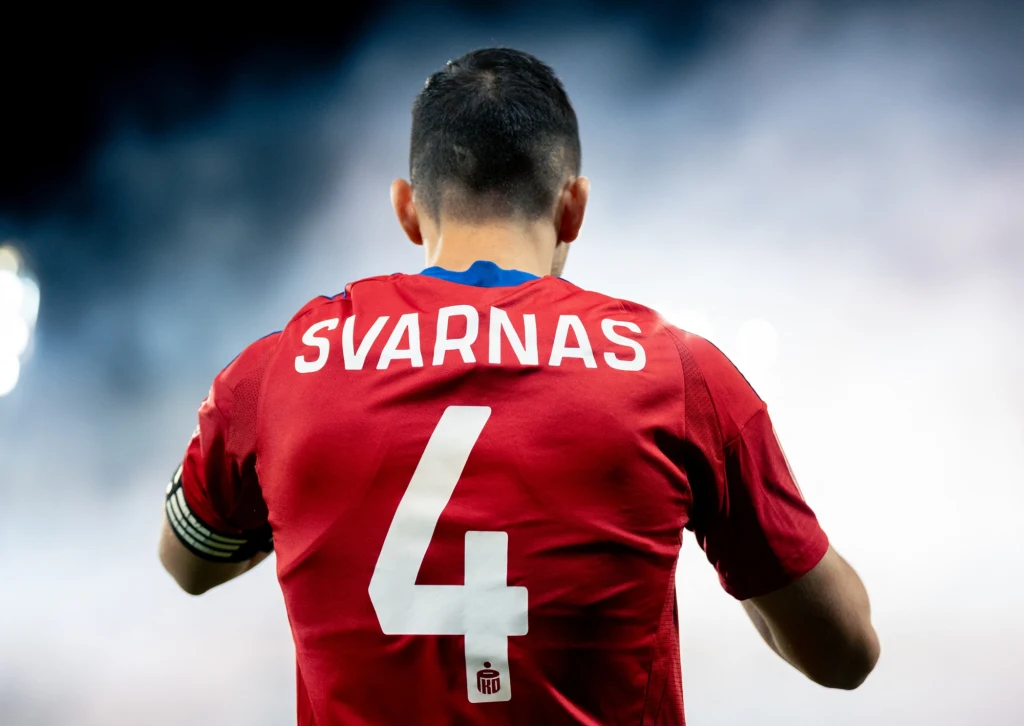 Stratos Svarnas Piłkarz w czerwonej koszulce z numerem 4 oraz napisem SVARNAS na plecach, widoczny od tyłu, w atmosferze stadionowej na tle jasnego światła i lekkiej mgły.