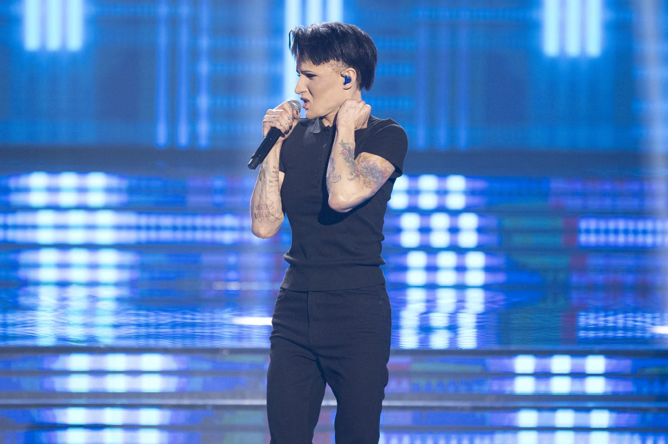 Kamil Studnicki jako Agnieszka Chylińska Osoba ubrana na czarno śpiewa na scenie, trzymając mikrofon i mając w uchu odsłuch, w tle widać kolorowe światła sceniczne oraz rozświetlone schody.