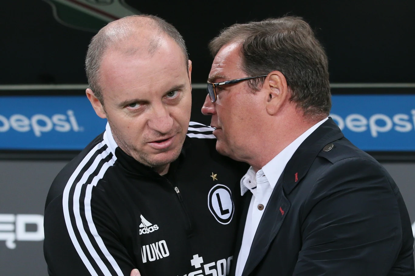 Dwóch mężczyzn w sportowych ubraniach znajduje się blisko siebie, jeden z nich szeptem przekazuje coś drugiemu. W tle widoczny fragment logo stadionowego baneru.