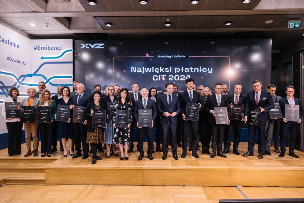 mBank Grupa osób ubranych w eleganckie stroje stoi na scenie, trzymając tablice z napisami. W tle znajduje się duży ekran z napisem największi płatnicy CIT 2024 w nowoczesnym wnętrzu. Panuje atmosfera oficjalnego wydarzenia.