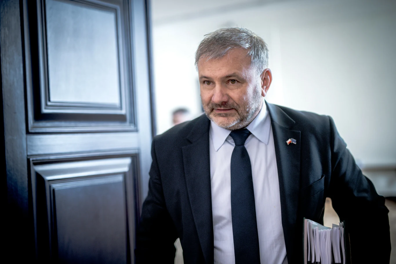 Mężczyzna w garniturze z dokumentami w ręku wchodzi do pomieszczenia przez otwarte drzwi, ma poważny wyraz twarzy, w tle rozmyte jasne ściany.