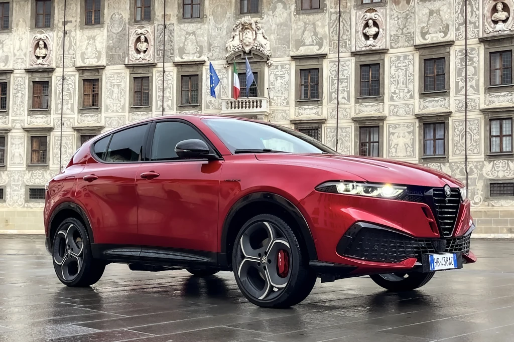 Czerwony samochód Alfa Romeo Tonale zaparkowany na tle zabytkowego budynku o bogato zdobionej fasadzie, z widocznymi flagami przed wejściem.