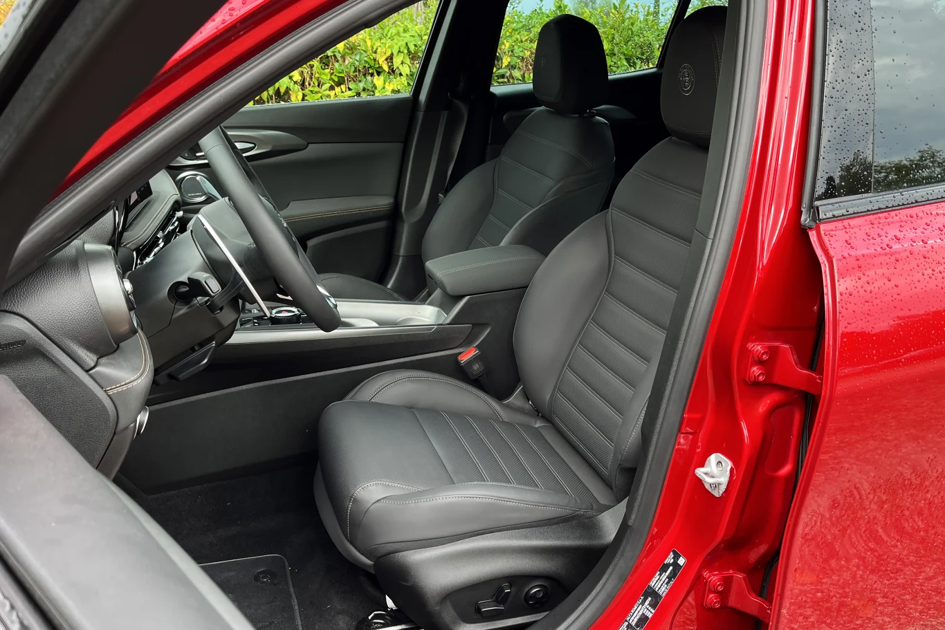 Czarne, skórzane fotele z przodu w nowoczesnym, czerwonym samochodzie, widoczne szczegóły wnętrza jak kierownica, deska rozdzielcza oraz boczek drzwi.