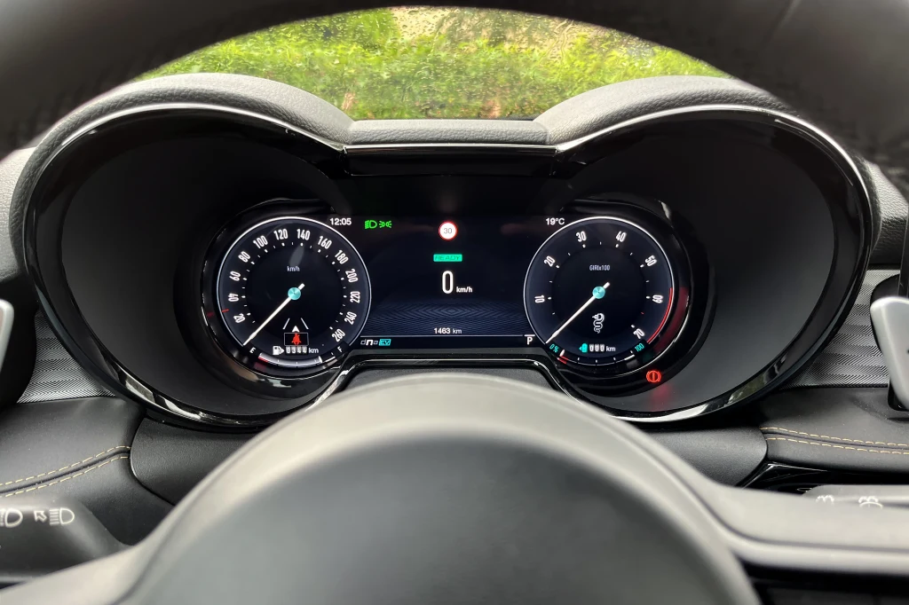 Cyfrowy panel wskaźników samochodu z prędkościomierzem, obrotomierzem i wyświetlaczem centralnym pokazującym aktualną prędkość, poziom paliwa oraz inne dane eksploatacyjne, otoczony elementami kierownicy.