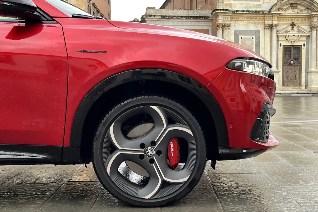 Czerwony samochód z nowoczesnymi felgami aluminiowymi zaparkowany na brukowanej powierzchni, na tle zabytkowego budynku z bogato zdobioną fasadą.