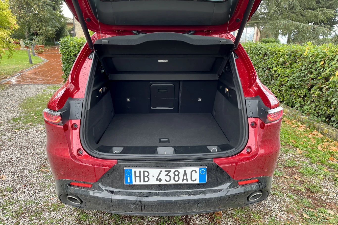 Otwarty bagażnik czerwonego samochodu osobowego zaparkowanego na żwirowej nawierzchni, w tle widoczna zielona roślinność i fragment domu.