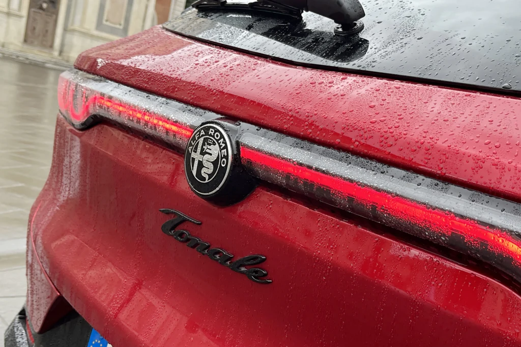 Czerwona klapa bagażnika samochodu Alfa Romeo Tonale z charakterystycznym emblematem marki oraz napisami, pokryta kroplami deszczu, widoczne tylne światła LED.