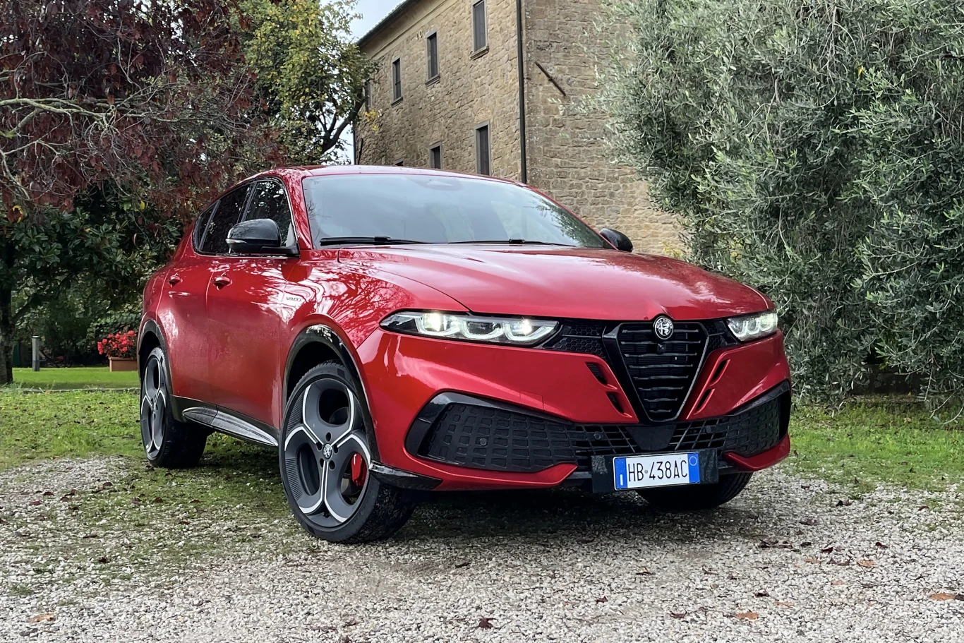 Czerwony samochód Alfa Romeo Tonale zaparkowany na żwirowym podjeździe, w otoczeniu zielonych drzew i starego budynku z kamienia. Wyraźnie widoczne sportowe linie nadwozia, charakterystyczny grill oraz aluminiowe felgi.