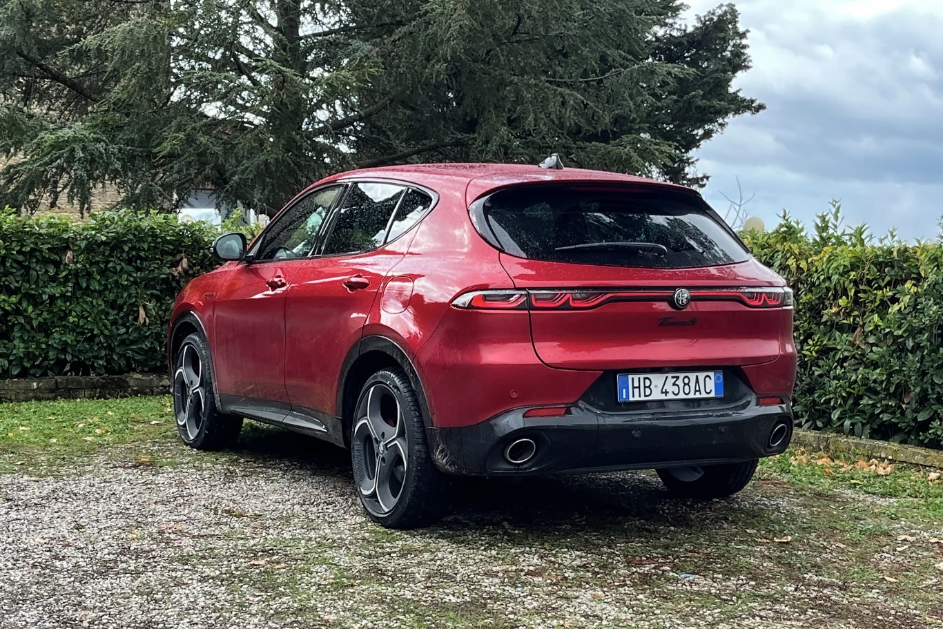 Czerwony samochód typu SUV marki Alfa Romeo zaparkowany na żwirowej nawierzchni, w otoczeniu zielonych krzewów i drzew. Widoczne tylne światła, tablica rejestracyjna oraz masywne koła.