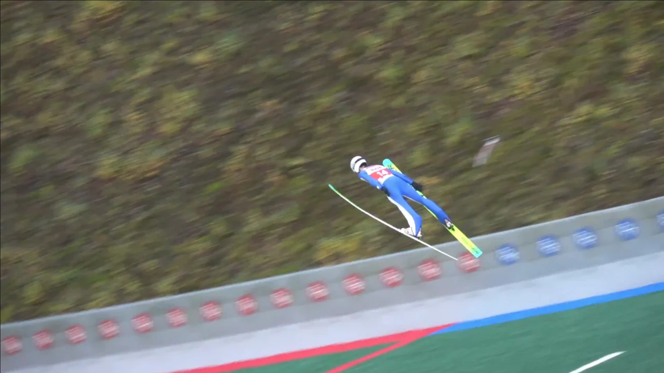 Kacper Tomasiak w locie Zawodnik w niebieskim stroju wykonuje skok narciarski na zielonym zeskoku, w tle widać fragment stoku porośnięty trawą oraz część infrastruktury skoczni.