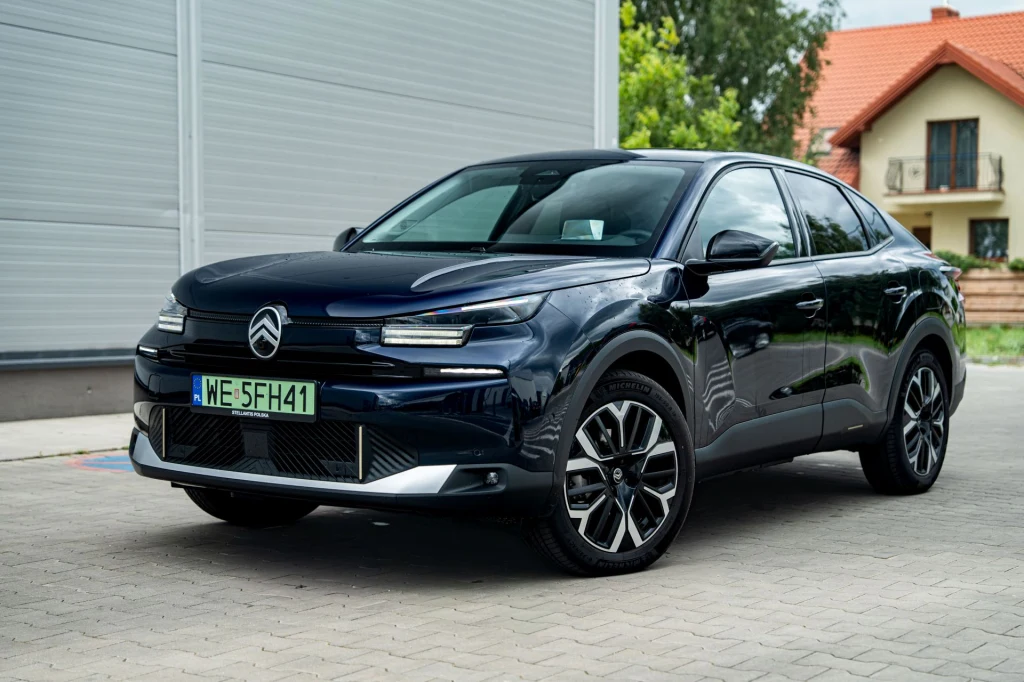 Czarny samochód Citroën C4 stoi zaparkowany na brukowanej nawierzchni obok jasnego budynku z garażem, w tle widoczne drzewa i dom z czerwoną dachówką.