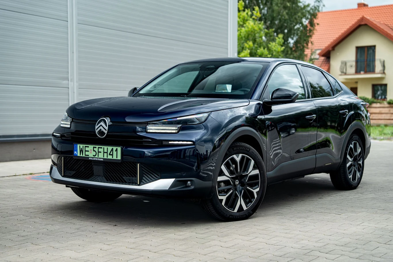 Czarny samochód Citroën C4 stoi zaparkowany na brukowanej nawierzchni obok jasnego budynku z garażem, w tle widoczne drzewa i dom z czerwoną dachówką.