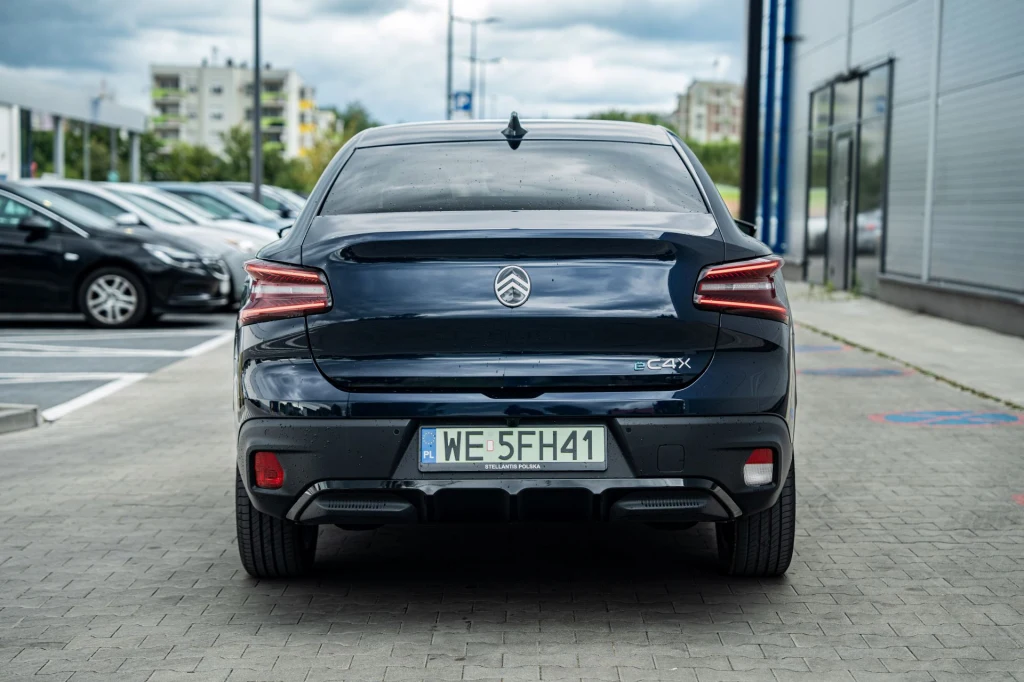 Czarny samochód Citroën C4 X zaparkowany tyłem na tle nowoczesnego budynku, widoczna rejestracja oraz tylne światła, w tle inne zaparkowane auta i bloki mieszkalne.