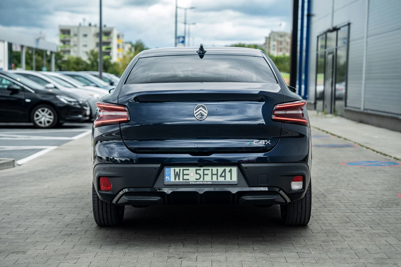 Czarny samochód Citroën C4 X zaparkowany tyłem na tle nowoczesnego budynku, widoczna rejestracja oraz tylne światła, w tle inne zaparkowane auta i bloki mieszkalne.