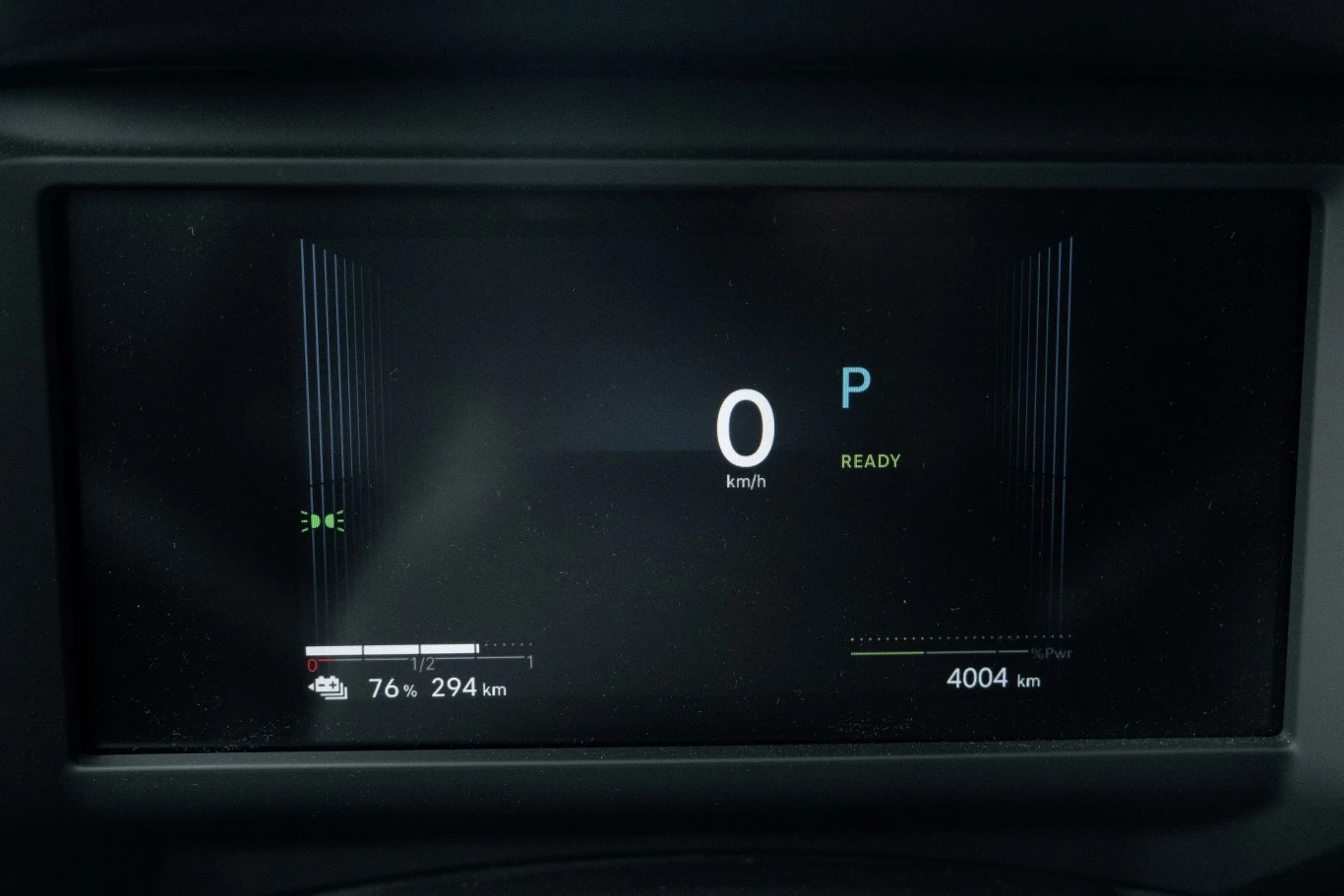 Panel cyfrowy samochodu elektrycznego z wyświetlonymi informacjami takimi jak prędkość 0 km/h, tryb P, poziom naładowania baterii 76% oraz przebieg 4004 km.