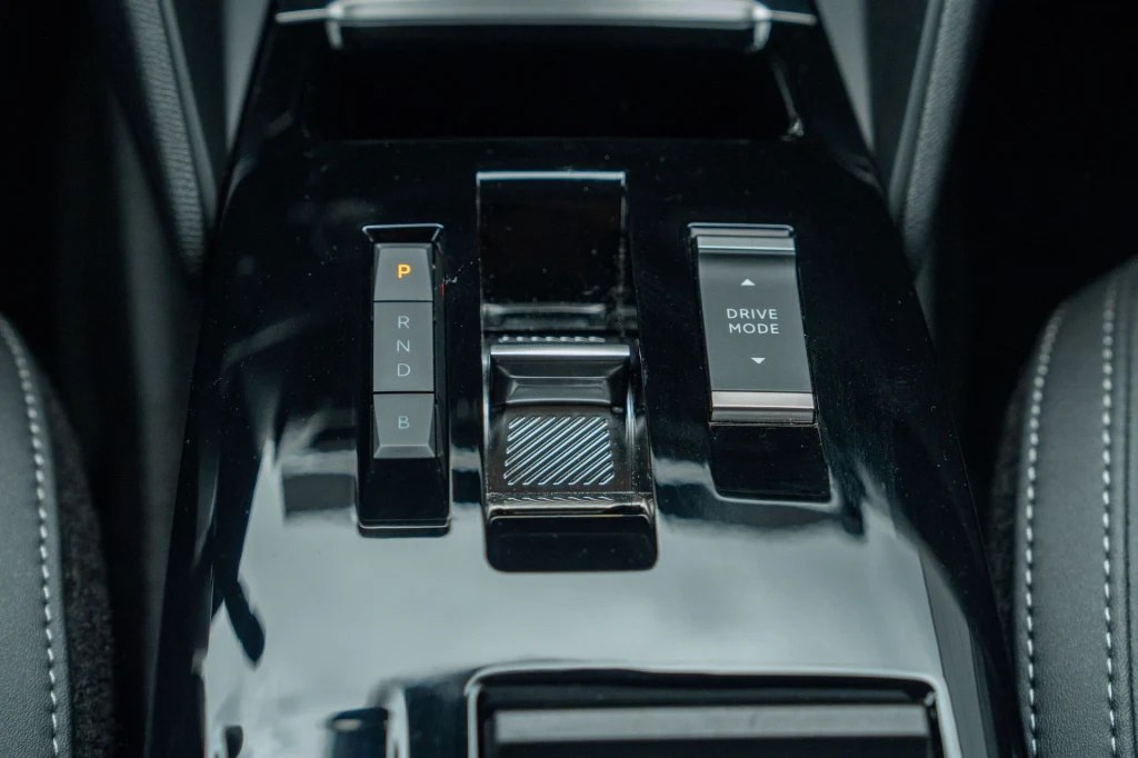 Czarny, błyszczący panel sterowania w samochodzie z wyraźnymi przyciskami do zmiany biegów oraz przyciskiem do wyboru trybu jazdy.