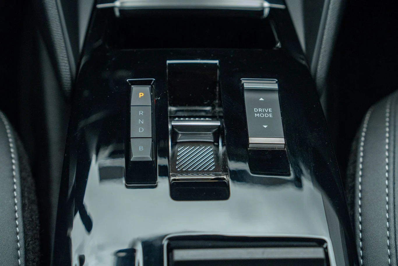 Czarny, błyszczący panel sterowania w samochodzie z wyraźnymi przyciskami do zmiany biegów oraz przyciskiem do wyboru trybu jazdy.