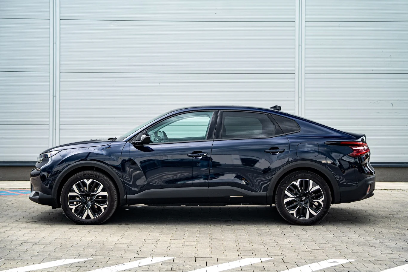 Czarny samochód osobowy o nowoczesnej sylwetce, stojący na betonowej nawierzchni na tle jasnej metalowej ściany. Wyróżniają się duże koła oraz sportowa linia nadwozia.