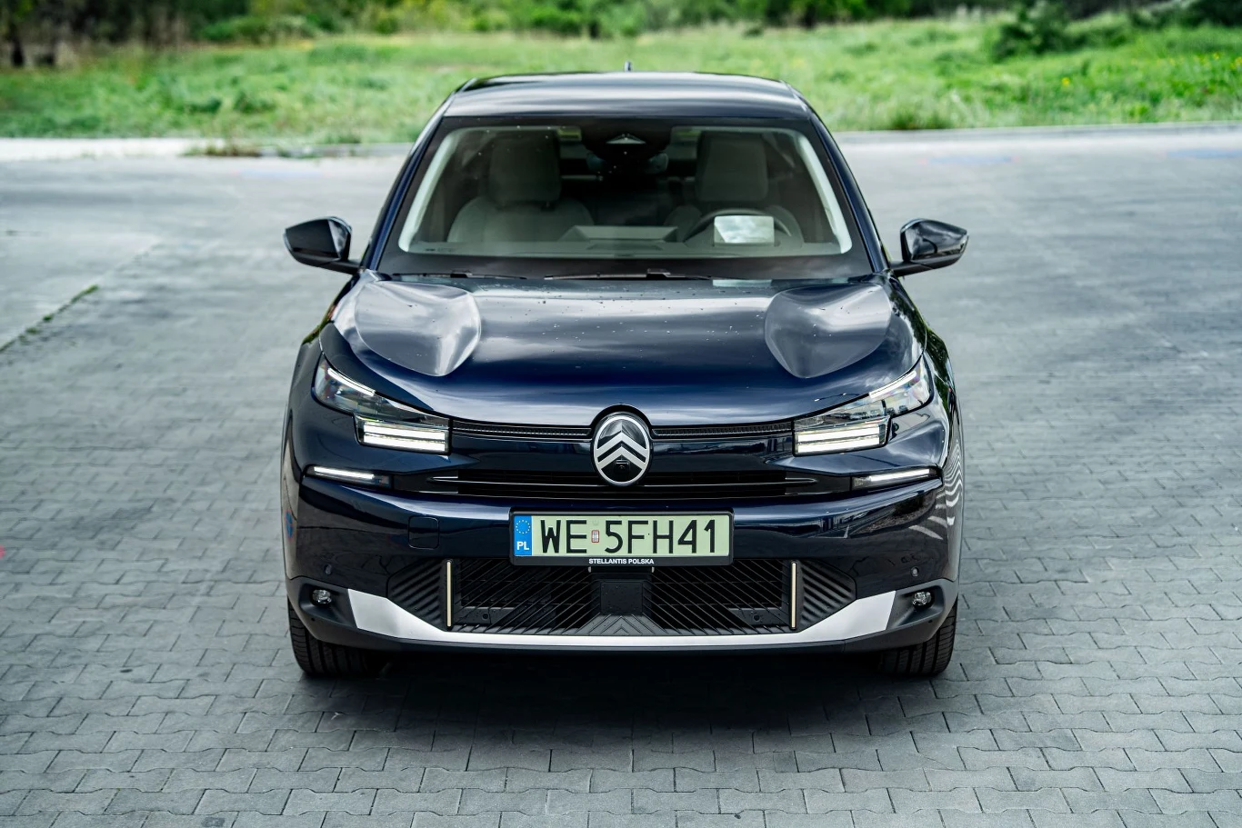 Czarny samochód Citroën stoi zaparkowany przodem na jasnej, brukowanej nawierzchni. Widoczna tablica rejestracyjna WE 5FH41. W tle rozmyta zieleń i szara ścieżka.