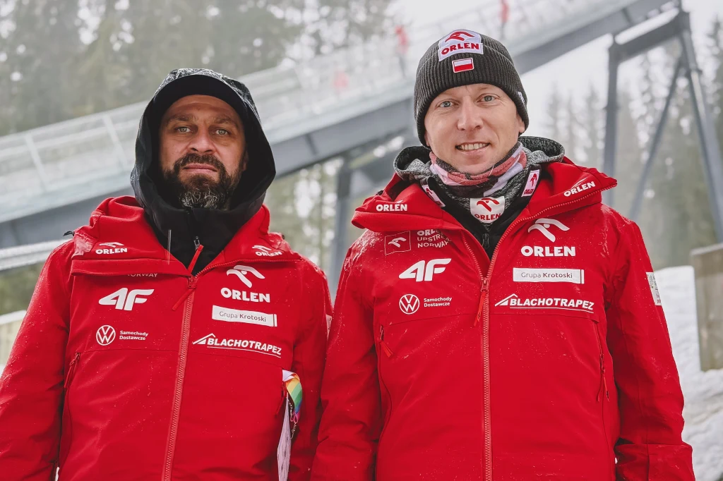 Marcin Bachleda (po lewej) i Stefan Hula - trenerzy kadry kobiet Dwóch mężczyzn w czerwonych kurtkach zimowych z logo sponsorów, stojących na tle skoczni narciarskiej w zimowej scenerii. Obaj są uśmiechnięci, mają czapki i szaliki, a w tle widać ośnieżone drzewa.