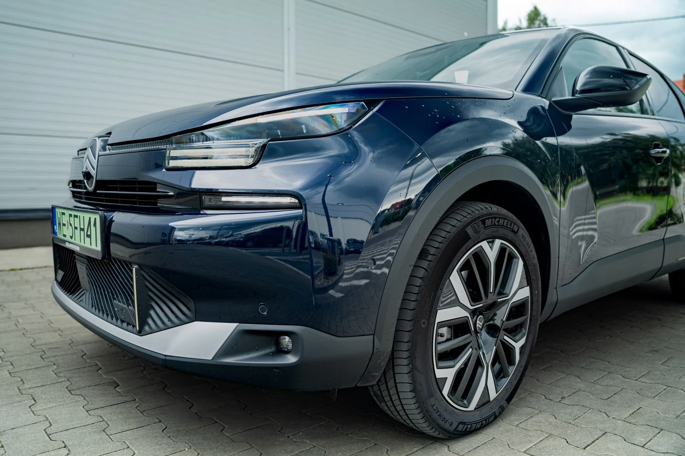 Nowoczesny samochód osobowy w ciemnoniebieskim kolorze zaparkowany przy budynku, widoczny z przodu z charakterystycznym grillem, reflektorami oraz aluminiowymi felgami.