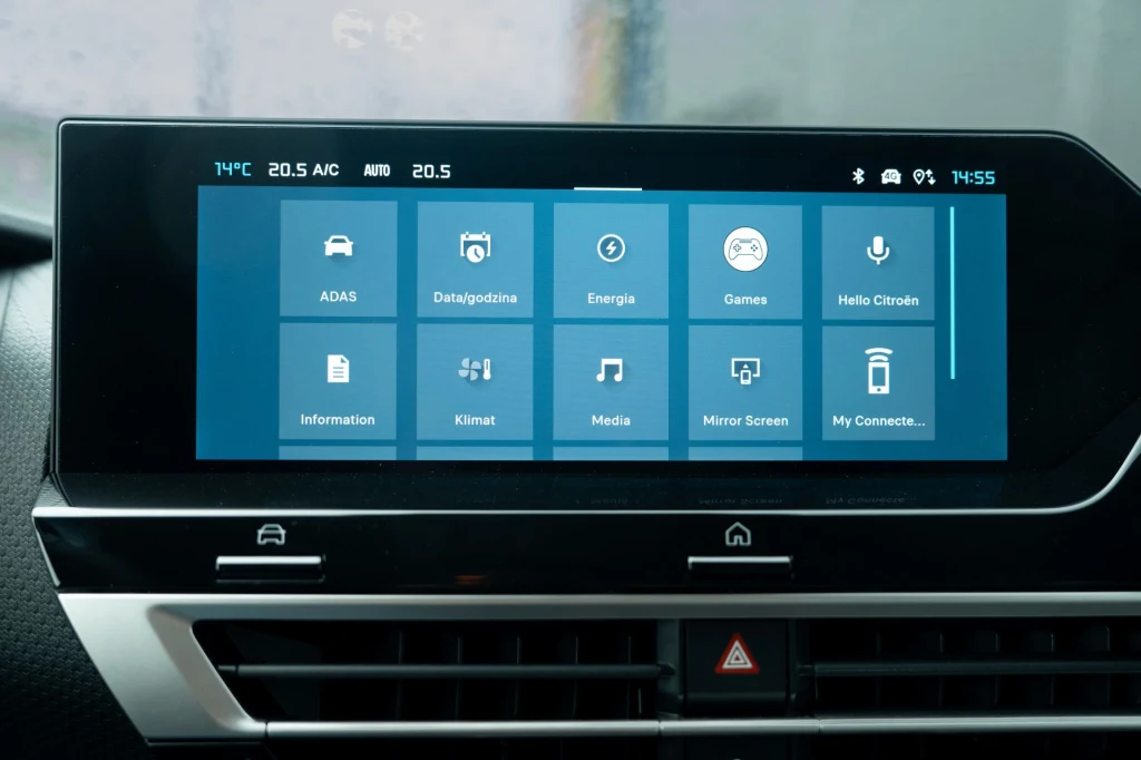 Dotykowy wyświetlacz systemu multimedialnego w samochodzie, wyświetlający główne ikony menu takie jak nawigacja, energia, gry, klimat, media i inne funkcje pojazdu. Poniżej ekran wywietrznik powietrza oraz przycisk awaryjnych świateł.