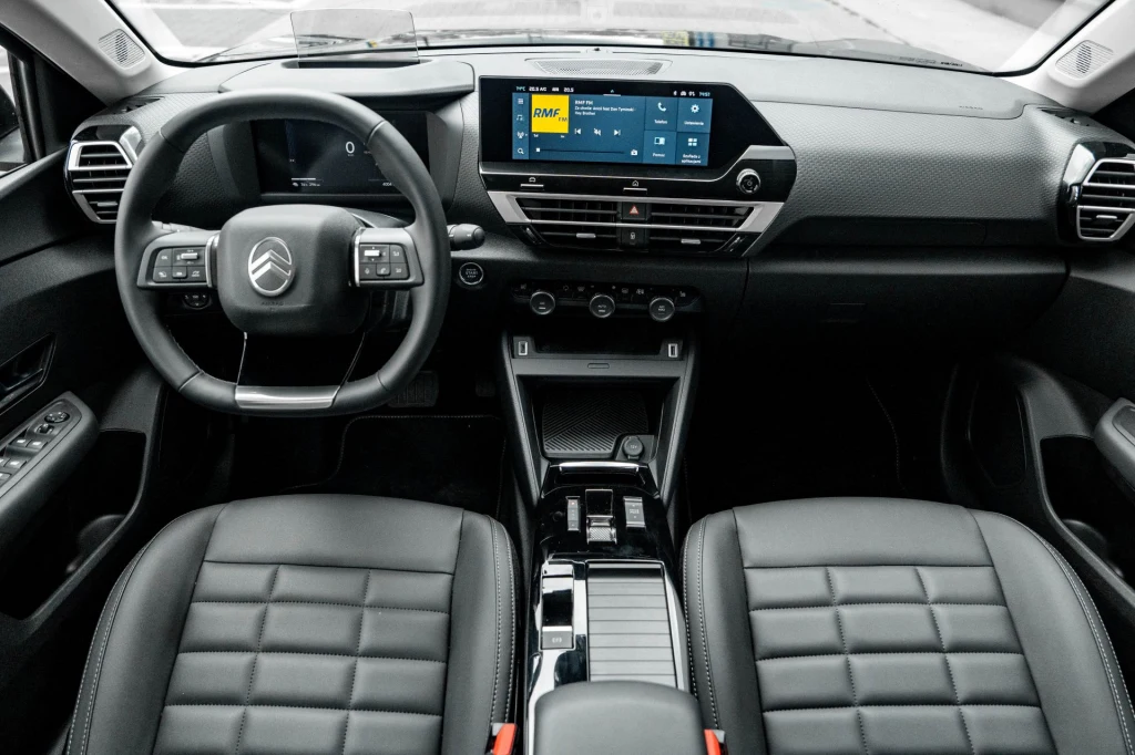 Nowoczesna deska rozdzielcza samochodu Citroën z dużym ekranem dotykowym, wielofunkcyjną kierownicą i dwoma wygodnymi fotelami z czarnej skóry. Wnętrze utrzymane w minimalistycznym stylu z dominacją czerni i srebrnych detali.
