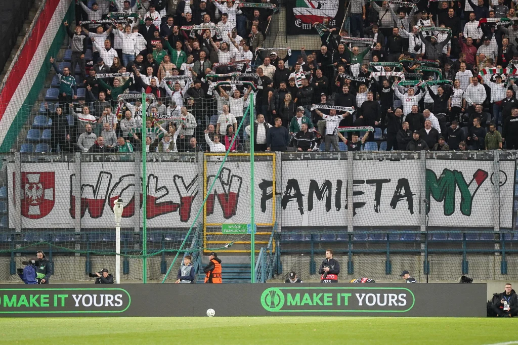 Transparent wywieszony przez kibiców Legii w trakcie meczu z Szachtarem Donieck Tłum kibiców na stadionie piłkarskim trzyma szaliki, za barierką widoczny duży baner z napisem „Wołyń pamiętamy” napisanym literami o odmiennych kolorach. Na banerze znajduje się również herb w biało-czerwonych barwach.