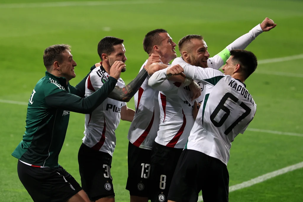 Rafał Augustyniak odbiera gratulacje od partnerów. Do siatki rywala trafił dwukrotnie Grupa piłkarzy Legii Warszawa w białych koszulkach w trakcie dynamicznej celebracji zdobytej bramki podczas meczu, jeden z zawodników unosi rękę w geście triumfu, pozostali przytulają się i uśmiechają.