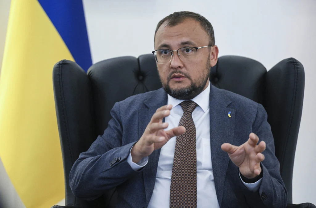 Wasyl Bodnar, ambasador Ukrainy w Polsce Mężczyzna w okularach i garniturze siedzi na dużym, ciemnym fotelu, wykonując gesty rękami, na tle częściowo widocznej ukraińskiej flagi.