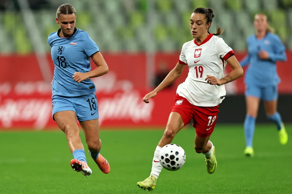 Dwie piłkarki w dynamicznym pojedynku o piłkę podczas meczu, zawodniczka w białej koszulce z numerem 19 reprezentuje Polskę, a zawodniczka w jasnoniebieskim stroju walczy o przejęcie futbolówki, w tle rozmyte trybuny stadionu.