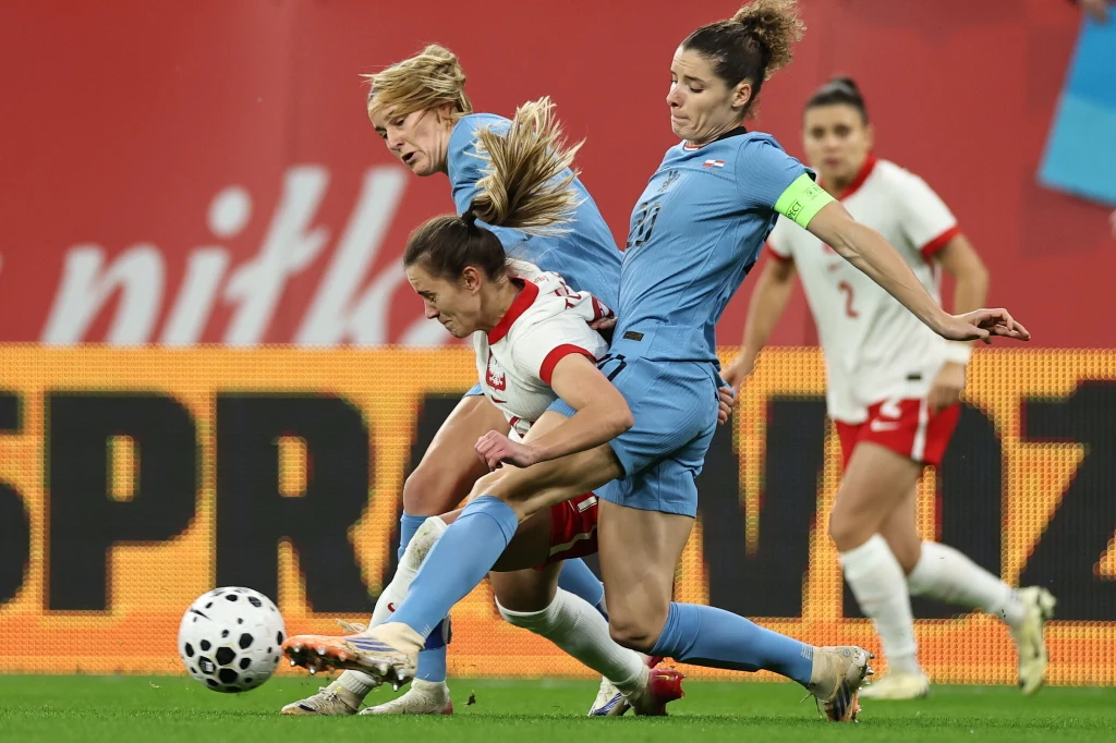 Dynamiczna walka o piłkę podczas meczu piłki nożnej kobiet, skupiająca się na trzech zawodniczkach w akcji, jedna z nich w jasnoniebieskim stroju, dwie w biało-czerwonych. W tle tablica reklamowa i inne zawodniczki obserwujące przebieg akcji.