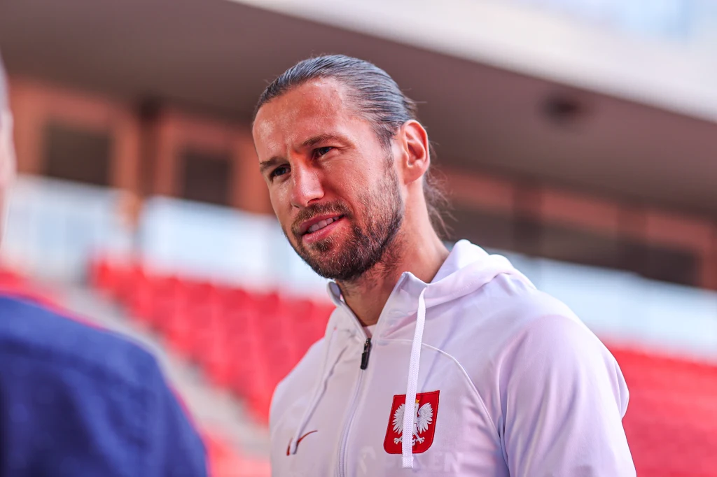 Grzegorz Krychowiak Mężczyzna o długich włosach spiętych z tyłu, ubrany w białą bluzę z godłem Polski, stoi na tle czerwonych trybun stadionowych i rozmawia z inną osobą.