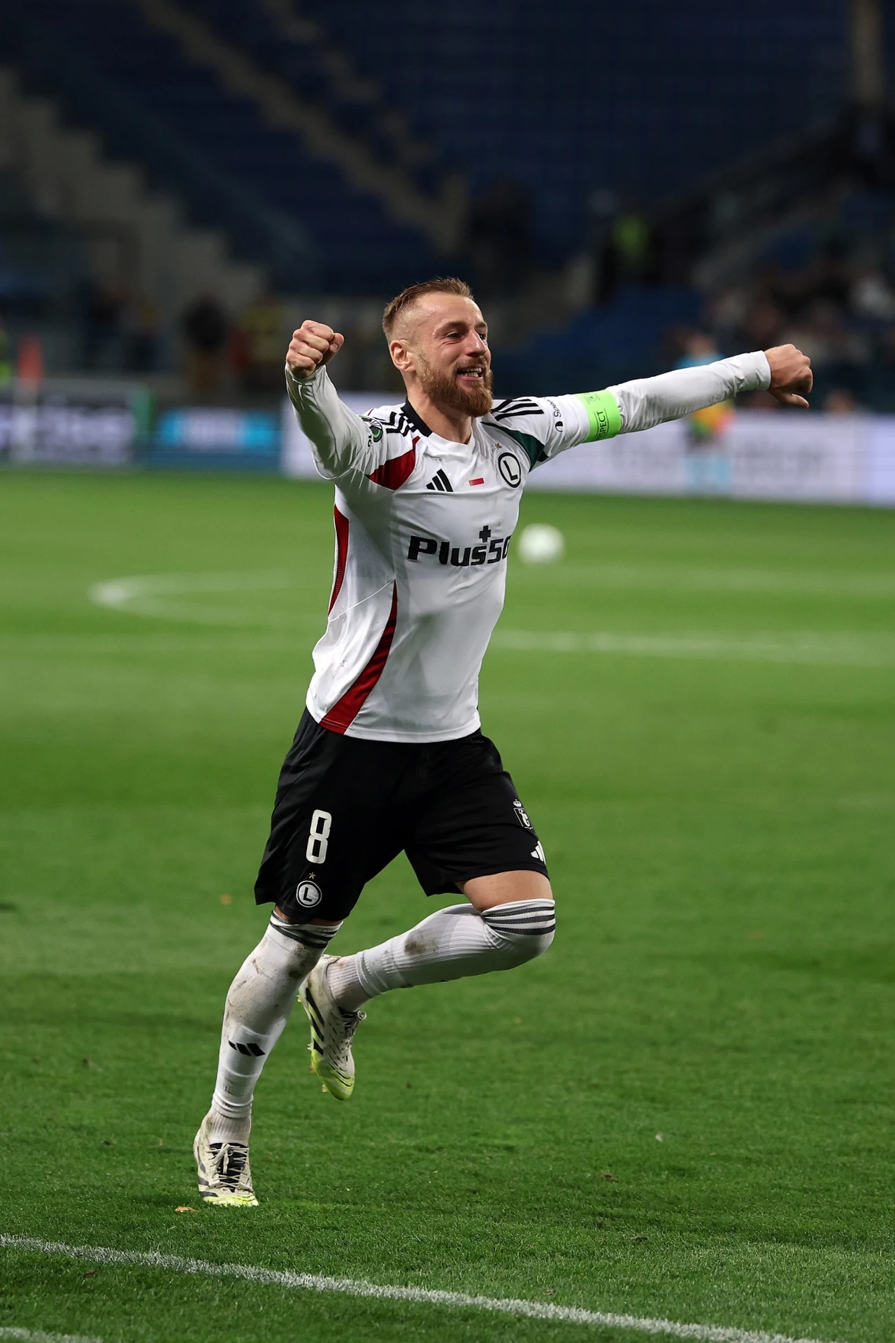 Rafał Augustyniak Piłkarz w białym stroju z numerem 8 i opaską kapitana biegnie po murawie stadionu, unosi ręce i wyraża radość po zdobytym golu, w tle widoczne są rozmyte trybuny i fragmenty boiska.