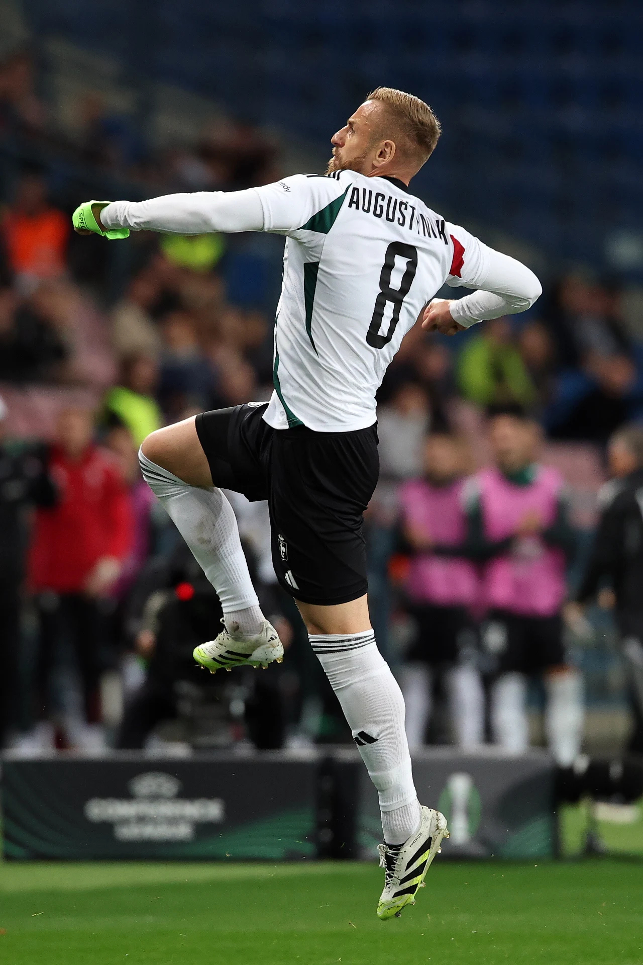 Rafał Augustyniak Piłkarz w białym stroju z numerem 8 i nazwiskiem Augustyniak na plecach wykonuje dynamiczny skok z zaciśniętą pięścią w uniesionej ręce, na tle stadionu z widocznymi kibicami i ławką rezerwowych.