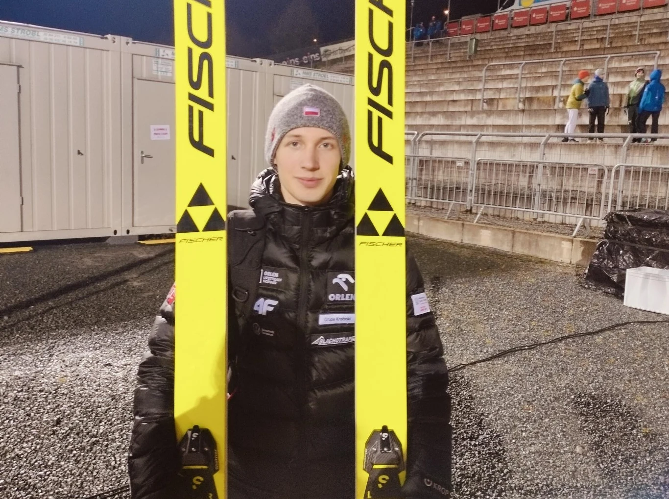 Kacper Tomasiak Młody mężczyzna w zimowej kurtce i czapce z biało-czerwoną flagą trzyma długie, żółte narty Fischer, stojąc na tle trybun i kontenerów technicznych stadionu sportowego.