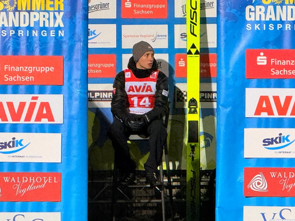 Zawodnik skoków narciarskich w sportowej odzieży i czapce siedzi na ławce w strefie przed startem, trzymając narty marki Fischer, w tle widoczne są liczne banery sponsorów i oznaczenia zawodów Summer Grand Prix.